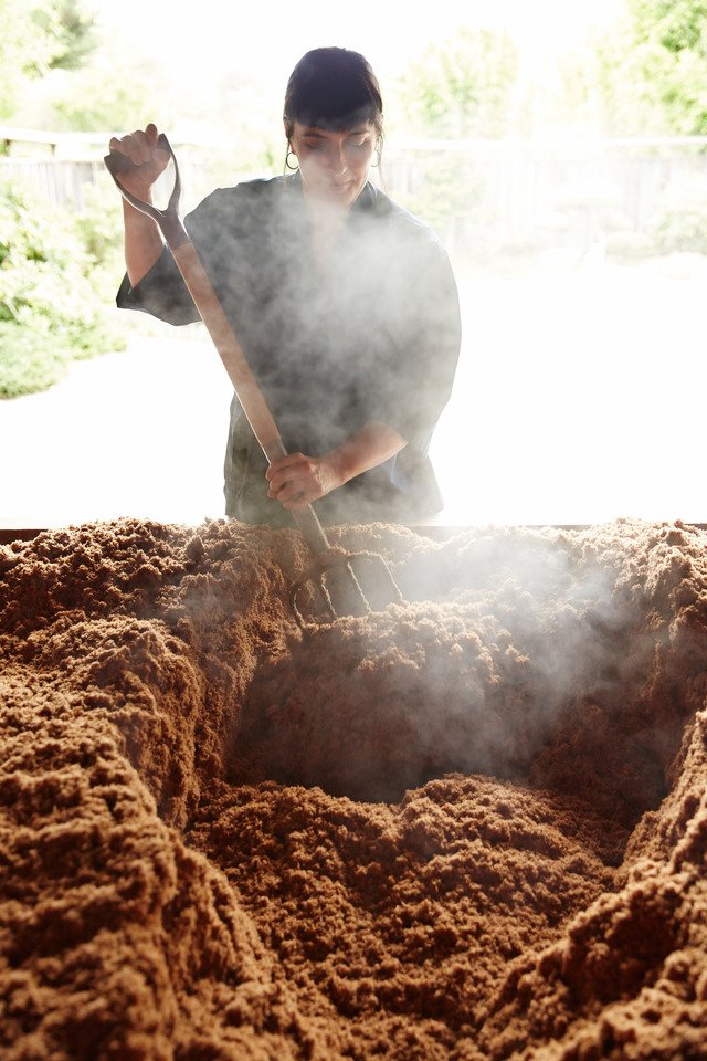 Person stirring steaming fermenting mixture outdoors.